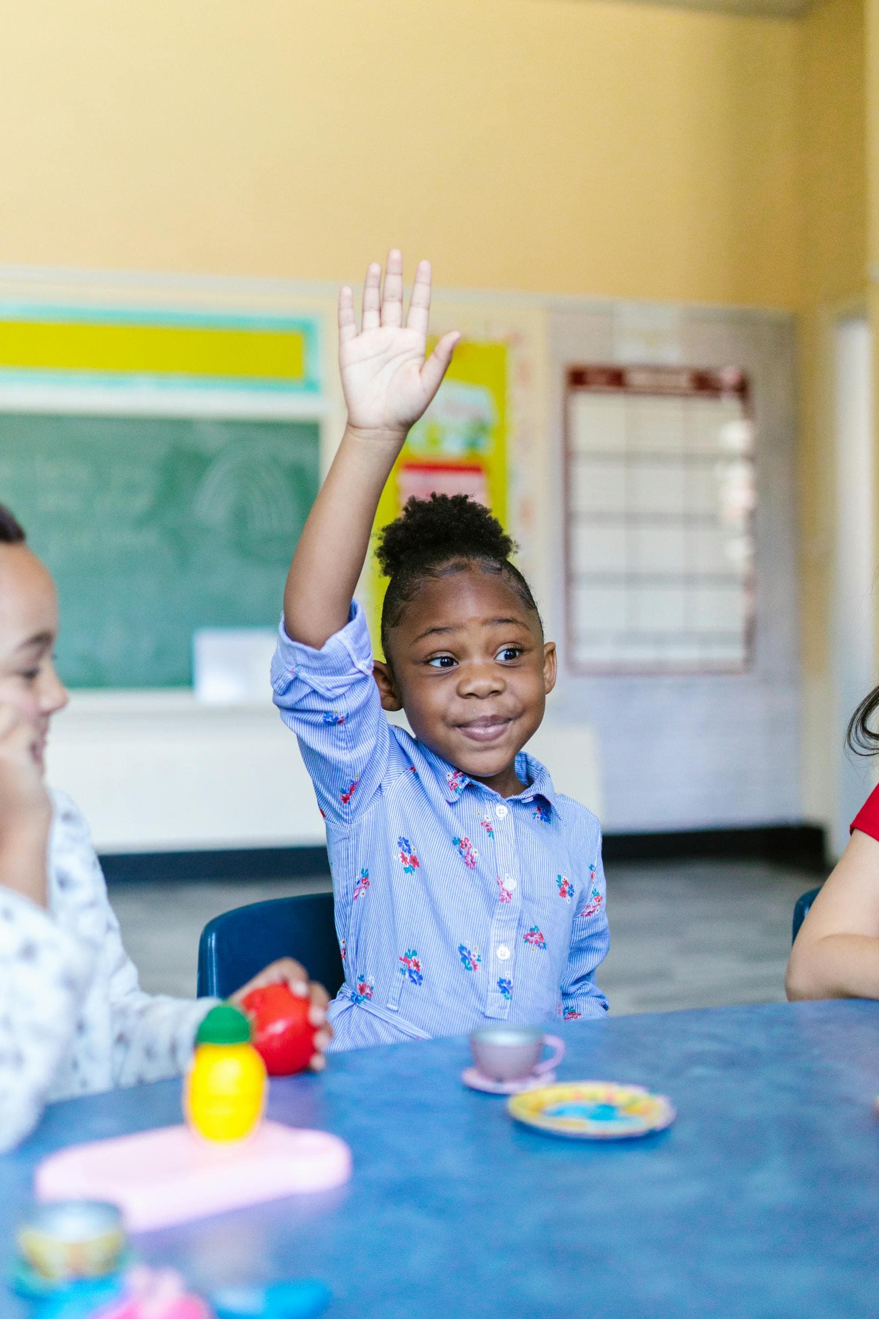 A young girl smiles and raises her hand in a bright preschool classroom.