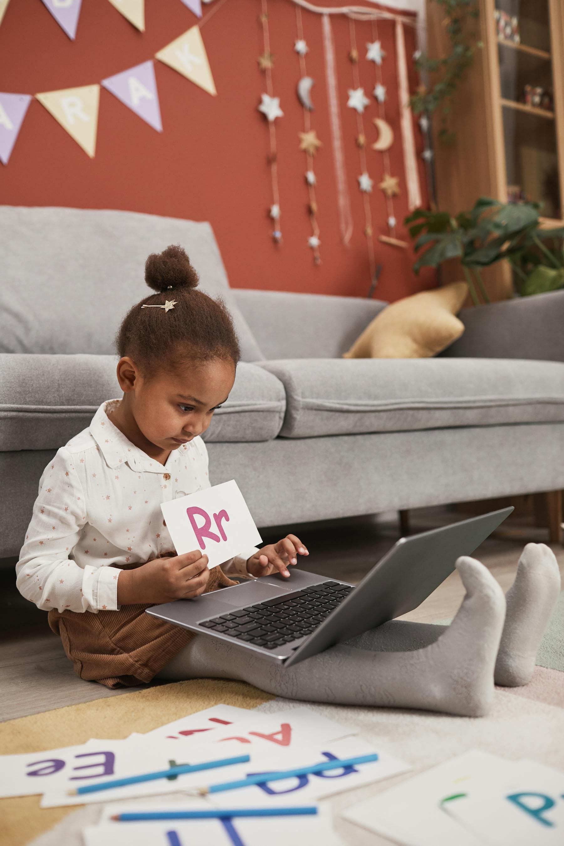 A young girl studies an alphabet card on the floor of a warm reading corner.